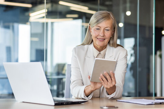Woman boss at workplace inside office, senior experienced gray-haired business woman uses tablet computer, happy checking financial data, banker finance woman with laptop.