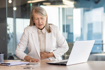 Fototapeta premium Sick overworked woman at workplace inside office has severe stomach pain, senior mature business woman holding hands on stomach massaging while sitting at desk with laptop.