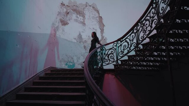 Halloween, a seductive witch in a black dress with gloves gracefully walks down the stairs of abandoned house, casting a shadow. The staircase is lit by eerie blue and red lamps. nighttime indoors.