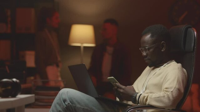 Medium shot of young Black male software developer using smartphone while sitting in office chair with laptop on his laps working at night office with diverse colleagues talking in background