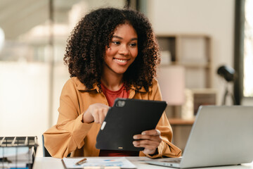 smiling African American university student with an afro hairstyle, holding a tablet and sitting at a desk with a laptop. online class learning from video conference