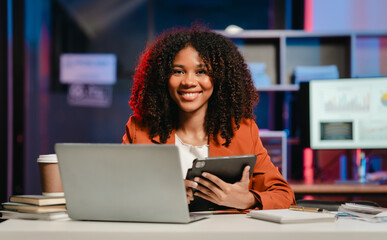 happy African American office worker with an afro, holding a tablet and working on a laptop at a...