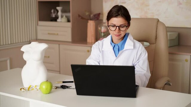 Female nutritionist working on laptop and writing diet plan for patient. Young beautiful smiling nutritionist consulting patient online at desk in wellness clinic. Right nutrition and diet concept