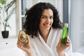 Smiling african woman in after-shower outfit presenting green avocado in fruit and cosmetic oil...