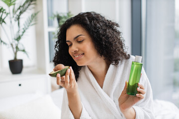 Beautiful calm female in bathrobe looking at camera with avocado half in hand while holding...