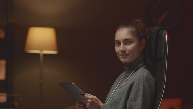 Waist up portrait of young female Caucasian IT coordinator with digital tablet smiling at camera while sitting in office chair at dark night office with red neon light