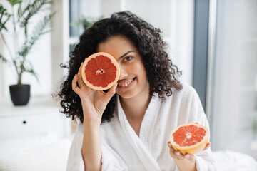 Attractive calm female in white bathrobe covering one eye with grapefruit half on blurred...