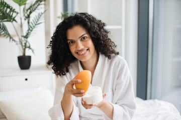 Happy young multinational woman in bathrobe holding half of grapefruit and making homemade cream...