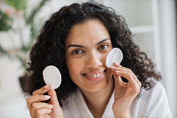 Portrait of young multiracial woman in white bathrobe holding cotton cosmetic sponges and smiling on camera on background of light bedroom. Natural beauty and skin care concept.