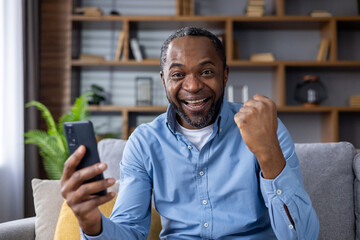 Portrait of a happy African American man sitting on the couch at home, using a mobile phone, and...
