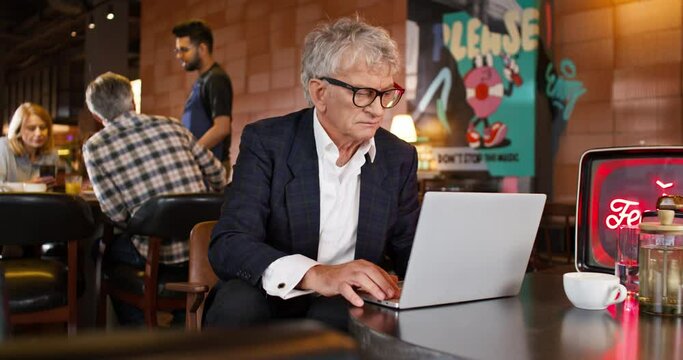 Mature businessman with elegant suit working on his portative laptop while sitting in cafe. In background customers communicating or making order with waiter. People resting in restaurant.