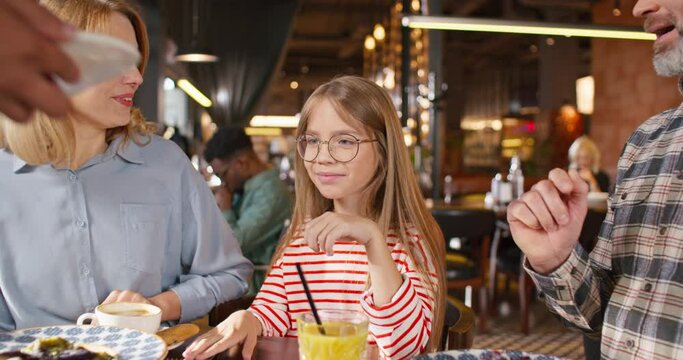 Happy Caucasian family resting together in cozy cafe. Hardworking waiter carrying tasty food for little girl. Satisfied people getting ready to eat food in local restaurant. Weekend concept.