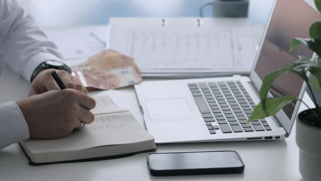 A young man writes financial expenses and income in a notebook. Control of banking operations. There is a laptop, a notebook, a folder with documents, a smartphone and cash on the table.