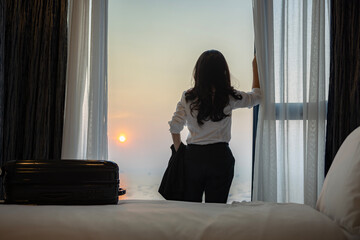 Businesswoman with suitcase opening the curtains and looking at the view from the window in luxury room of the expensive hotel after business meeting, Business trip, Booking hotel during your vacation
