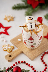 Winter hot chocolate with marshmallows in a mug with marshmallow snowman on a light background. Cozy still life with cocoa drink, cookies and decorations, gingerbread cookies, fir and poinsettia.