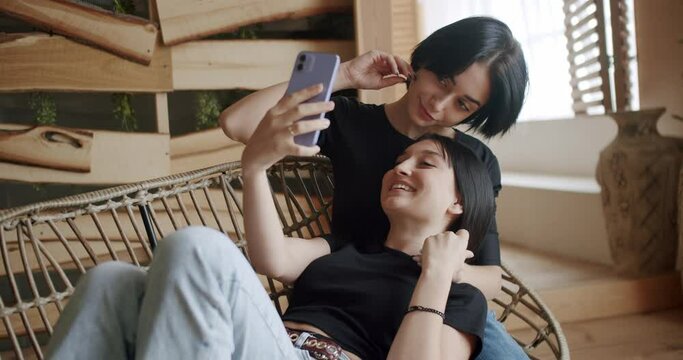 Two young woman lesbian spending time together, relaxing on armchair, smile and make selfie on smartphone. Happy female couple using cellphone to take picture