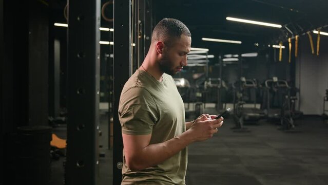 American Latino ethnic man browsing smartphone serious athlete fit sport guy personal fitness trainer instructor male in gym bodybuilder sportsman using mobile phone looking at camera happy smiling