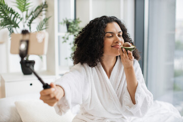 Beautiful multiracial female social media influencer in bathrobe streaming and introducing beauty...