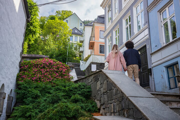 An interracial, heterosexual sexual couple holding hands, walks up a flight of stairs near the  Floibanen funicular during a summer morning. . - obrazy, fototapety, plakaty