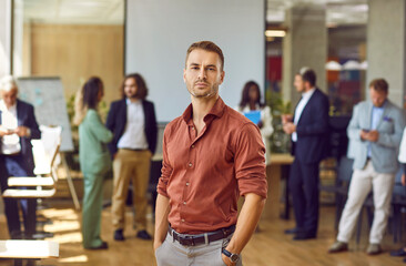 Fototapeta na wymiar Portrait of young businessman, corporate employee or team coach at professional business training event. Serious unsmiling handsome young man in brown shirt standing in office and looking at camera