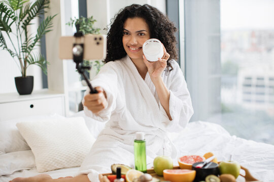 Beautiful young African woman social media influencer in bathrobe streaming and introducing beauty products to audience with phone holding moisturizing cream, skin care concept.