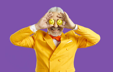 Portrait of funny optimistic senior man with happy emoji eyes. Studio shot of old guy wearing bright yellow jacket and orange bow tie covering his eyes with two smiley faces he's holding in his hands - obrazy, fototapety, plakaty