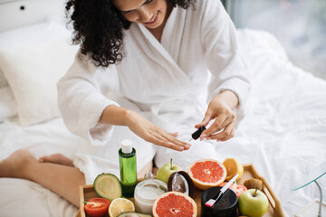 Top view of hands of multinational lady in dressing gown, paints nails while sitting near fruits and cosmetic containers on tray table on soft bed in bright room.