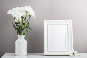 Empty white photo frame and a bouquet of white chrysanthemums, daisies in a ceramic vase. Postcard, home interior.