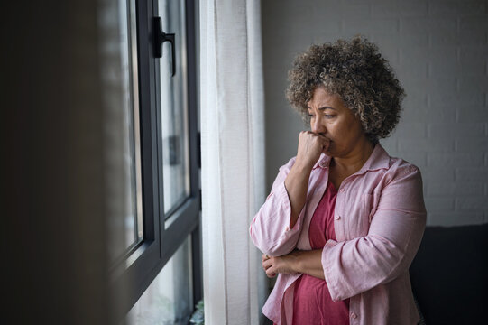 A mature woman of mixed race looking outside a large window, her expression reflecting sadness, concern, and a sense of depression