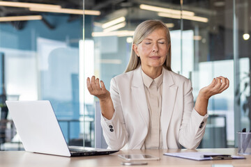 Businesswoman boss meditating with eyes closed at workplace inside office, senior woman working...