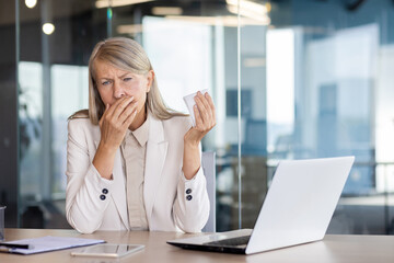 Senior gray-haired woman sick at workplace inside office, businesswoman boss sneezes has cold and...
