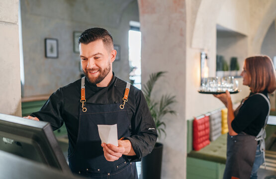Stylish bearded smiling waiter dressed black uniform using point of sale order terminal system touch Screen and waitress woman with tray. Successful people teamwork, restaurant industry concept image.