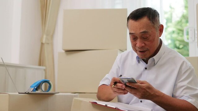 An elderly Asian man running an online business is checking information on his smartphone in preparation for sending items to customers. SME business. Entrepreneur. Working in reti
