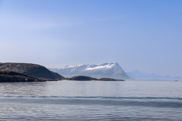 A picturesque summer scene from a ferry en route to Lofoten Island, Norway, showcasing the North Sea with serene waters and rugged mountains crowned with snow, under a sunny, azure sky