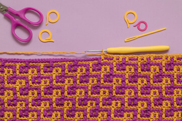 Handmade crochet texture with abstract pattern, crochet hook, scissors, knitting markers and yarns on a purple background. Copy space.
