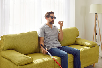 Portrait of a young bearded blind man in black eyeglasses holding cane and using digital assistant...