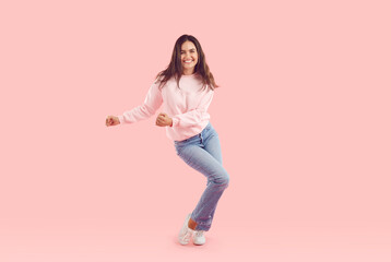 Fototapeta na wymiar Full body shot of happy, smiling woman in comfortable casual outfit. Portrait of cheerful beautiful young girl in pastel pink sweatshirt and blue jeans dancing on pink background in fashion studio