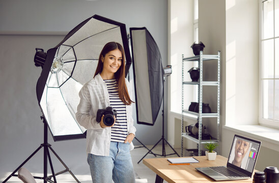 Young woman photographer in studio. Portrait of happy, smiling girl with photo camera standing in her creative workplace with laptop and lighting equipment. Professional photography concept