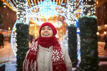 Joyful woman in knitted red cap and scarf walking on city square with festive colorful...