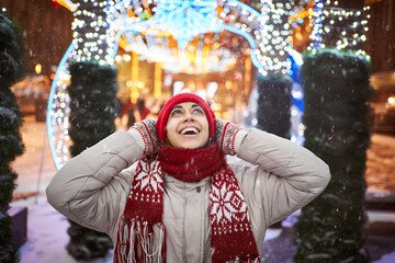 Happy laughing woman in red beanie and scarf walking on city square with festive colorful...