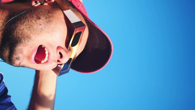 Vertical video of man wearing cap and solar eclipse glasses, looking sun with blue sky at background 