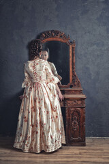 Beautiful woman in rococo style medieval dress standing near console table and looking at the mirror