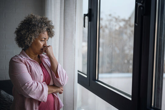 A mature woman of mixed race looking outside a large window, her expression reflecting sadness, concern, and a sense of depression