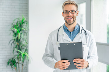 Handsome smiling medical doctor in white coat with clipboard, looking at camera and smiling while...