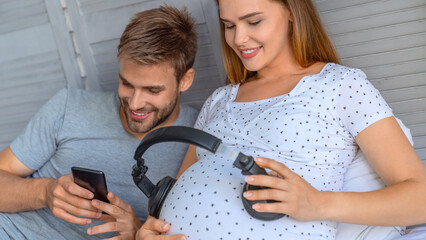 Laughing pregnant woman resting in sofa at home with earphones on naked belly while husband holding...