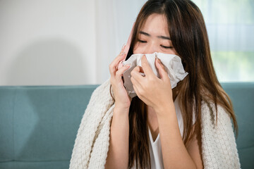 Asian young woman sick sitting down on sofa with cold blowing her nose hold tissues and talking on...