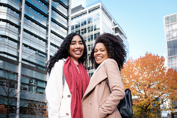 Smiling couple of female friends looking at camera surrounded by modern buildings.