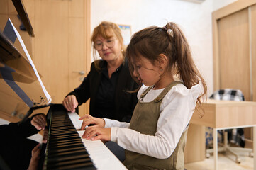 Kid girl playing piano, practicing music on chord instrument with her teacher during individual...