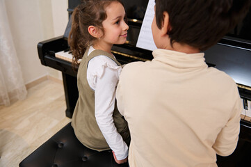 Rear view of two kids playing grand piano together indoor.