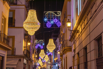 Christmas lights decoration in Sierpes street in the shape of a golden Empire style crystal candelabra, in the center city of Seville, Andalusia, Spain. Selective focus in the nearest line of lights - obrazy, fototapety, plakaty